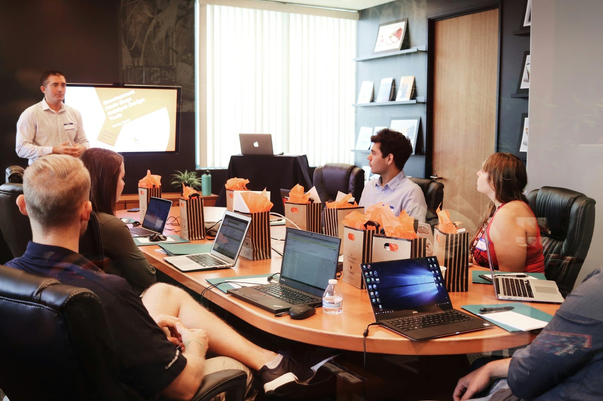A facilitator presents on a shared screen while a workshop group works on laptops around a conference table